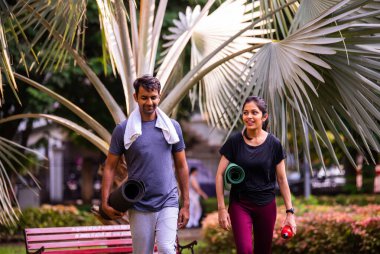 Hintli genç çift, yoga seansından sonra minderle birlikte parkta başarılı bir sabah antrenmanını kutluyor, refahın, zindeliğin ve yeşil havanın tadını çıkarıyor.