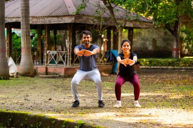 Hintli güzel genç çift güneşli bir günde parkın dışında birlikte çömeliyorlar. Günlük fitness rutinlerinin bir parçası olarak, güç, sağlık, motivasyon ve aktif sağlıklı yaşam tarzı hedeflerine odaklanıyorlar.