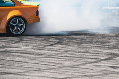 Arka tekerlekten çıkan beyaz dumanla birlikte gri hız pistinde sürüklenen sarı spor arabanın yan görüntüsü ve fotokopi alanı mevcut.
