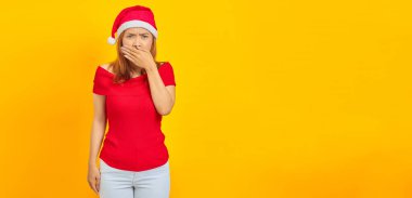 Şaşırmış genç Asyalı kadın Noel şapkası takıyor ağzını örtüyor sarı arka planda elleri var