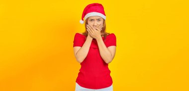 Şaşırmış genç Asyalı kadın Noel şapkası takıyor ağzını örtüyor sarı arka planda elleri var