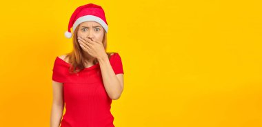 Şaşırmış genç Asyalı kadın Noel şapkası takıyor ağzını örtüyor sarı arka planda elleri var
