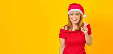 Gülümseyen Asyalı genç bir kadın Noel şapkası takıyor parmaklarını havaya kaldırıyor ve sarı arka plan hakkında bir fikri var.