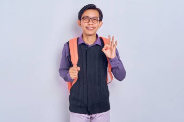 Sırt çantasıyla kolejden gülümseyen genç Asyalı erkek öğrenci gri arka planda izole edilmiş iyi bir hareket gösteriyor. Lise üniversite konsepti eğitimi