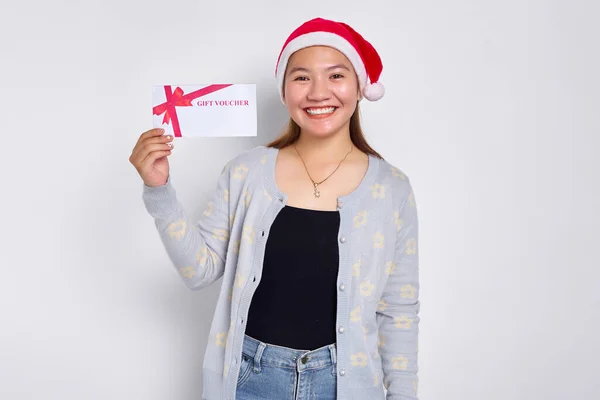 Smiling cheerful young Asian woman 20s in a Santa Claus hat Showing gift certificate coupon voucher card for the store, looking camera isolated on white studio background. Merry Christmas Concept