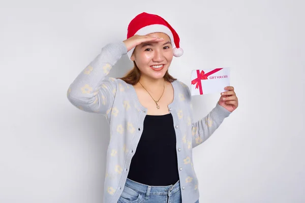Excited young Asian woman 20s in a Santa Claus hat holding a gift certificate coupon voucher card, looking far away distance isolated on white studio background. Merry Christmas Concept