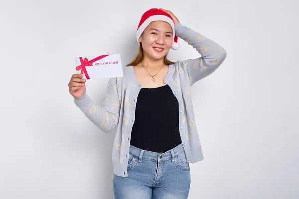 Thinking and pensive young Asian woman in a Santa Claus hat holding a gift certificate coupon voucher card, touching her head with hand isolated on white studio background. Merry Christmas Concept