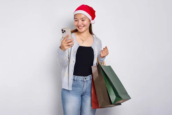 stock image Cheerful 20s young Asian woman wearing Christmas hat carrying shopping bags and looking for Christmas deals online on a mobile phone isolated on white background