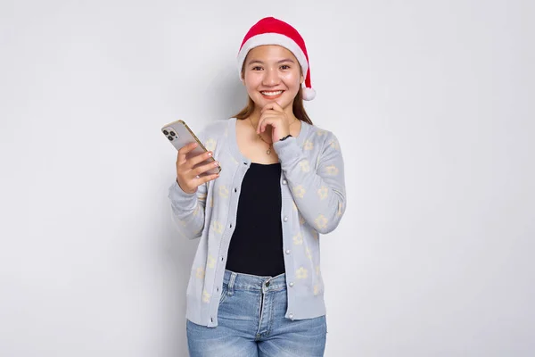 Uśmiechnięta Młoda Azjatka Świątecznym Kapeluszu Trzymająca Telefon Komórkowy Dotykająca Podbródka Obrazek Stockowy