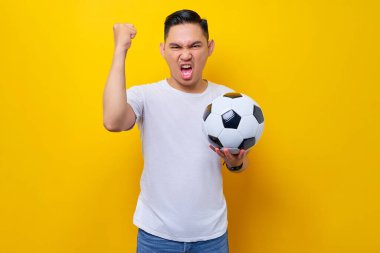 Taraftarlar futbol takımını destekleyeceği için heyecanlı. 20 'li yaşlarda Asyalı bir adam elinde futbol topu olan beyaz tişört giyer ve televizyonda canlı yayında sarı arka planda izole edilmiş bir jest yapar.