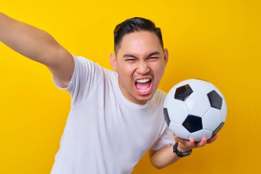 Beyaz tişörtlü, futbol topu taşıyan ve sarı arka planda izole edilmiş bir cep telefonunda selfie çeken Asyalı genç futbolcu hayranı heyecanlı. İnsanlar eğlence yaşam tarzı konsepti ile spor yapıyor.