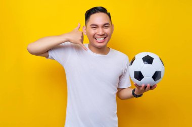 Asyalı genç bir futbol fanatiği elinde futbol topu tutan beyaz bir tişört giyiyor ve jestler gösteriyor sarı arka planda beni izole ediyor. İnsanlar eğlence yaşam tarzı konsepti ile spor yapıyor.