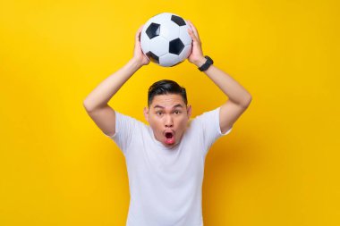 Beyaz tişörtlü neşeli Asyalı genç adam futbol fanatiği sarı arka planda izole edilmiş futbol topu hareketini yakalayan favori takımı neşelendiriyor. İnsanlar eğlence yaşam tarzı konsepti ile spor yapıyor.