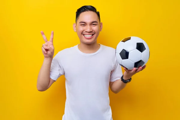 Smiling young Asian man football fan in a white t-shirt holding a soccer ball, showing a victory sign isolated on yellow background. People sport leisure lifestyle concept