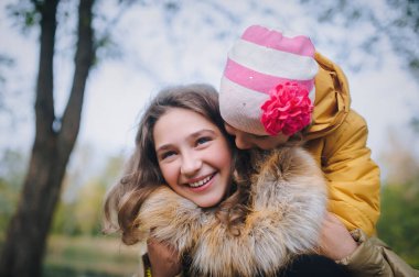 Küçük kız kardeş ablasını öpüyor. Mutlu gülümseyen çocuklar sonbahar parkında eğleniyor..