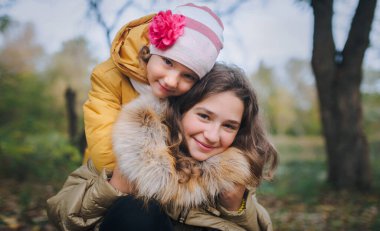 Mutlu gülümseyen çocuklar sonbahar parkında eğleniyorlar. Küçük kız kardeşi ablasına sarılıyor..