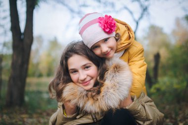 Küçük kız kardeş ablasına sarılıyor. Mutlu gülümseyen çocuklar sonbahar parkında eğleniyor..