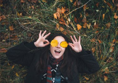 Kız, sonbahar ormanında sarı yaprakların üzerinde çimenlerin üzerinde uzanır ve şakayla gözlerini sarı yapraklarla kapar..