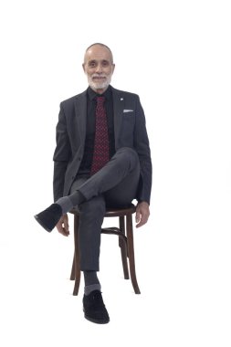 Önde oturan bir adam, takım elbiseli, kravatlı, çapraz bacaklı beyaz arka plandaki kameraya bakıyor.