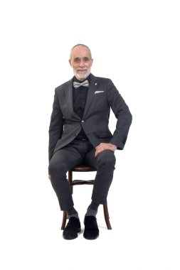 Önde oturan bir adam, takım elbiseli ve papyonlu beyaz arka planda kameraya bakıyor.