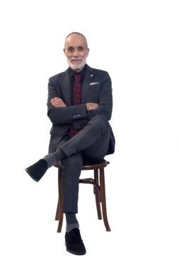 Önde oturan bir adam, takım elbiseli, kravatlı, çapraz bacaklı beyaz arka plandaki kameraya bakıyor.