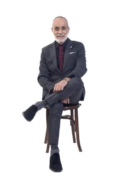 Önde oturan, takım elbiseli, kravatlı, çapraz bacaklı beyaz arka planda kameraya bakan bir adam var.