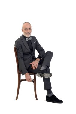 Takım elbiseli ve papyonlu bir adam kameraya bakıyor ve beyaz arka planda bacak bacak üstüne atıyor.