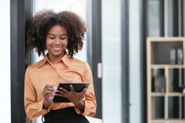 Mutlu Afrikalı iş kadını dijital aletlerle sohbet ediyor, cep telefonu tutuyor, dizüstü bilgisayar kullanıyor, kameraya bakıyor, gülümsüyor. Ev işyerindeki serbest çalışan kafasından vurulmuş. Yüksek kalite