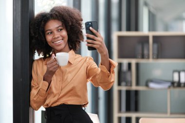 Mutlu Afrikalı iş kadını dijital aletlerle sohbet ediyor, cep telefonu tutuyor, dizüstü bilgisayar kullanıyor, kameraya bakıyor, gülümsüyor. Ev işyerindeki serbest çalışan kafasından vurulmuş. Yüksek kalite