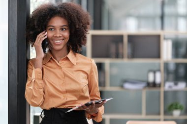 Mutlu Afrikalı iş kadını dijital aletlerle sohbet ediyor, cep telefonu tutuyor, dizüstü bilgisayar kullanıyor, kameraya bakıyor, gülümsüyor. Ev işyerindeki serbest çalışan kafasından vurulmuş. Yüksek kalite