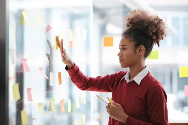 stock image Close up of focused african American female employee write down on colorful sticky notes manage list, concentrated biracial woman work on startup brainstorm collaborate plan on stickers on glass wall