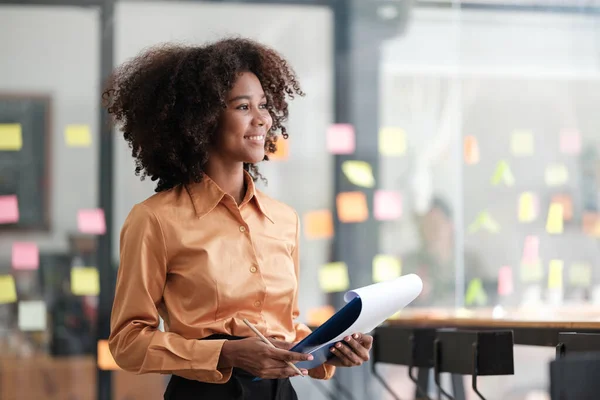 Close up of focused african American female employee write down on colorful sticky notes manage list, concentrated biracial woman work on startup brainstorm collaborate plan on stickers on glass wall