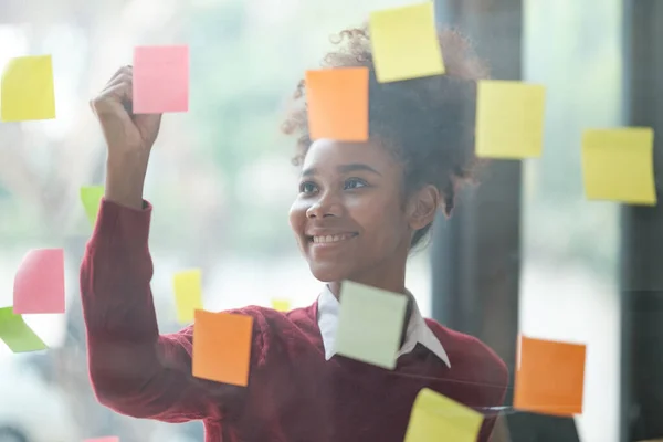 Close up of focused african American female employee write down on colorful sticky notes manage list, concentrated biracial woman work on startup brainstorm collaborate plan on stickers on glass wall