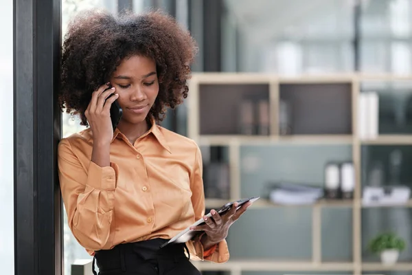 stock image Happy African business woman chatting on digital gadgets online, holding mobile phone, using laptop computer, looking at camera, smiling. Freelance employee at home workplace head shot. High quality