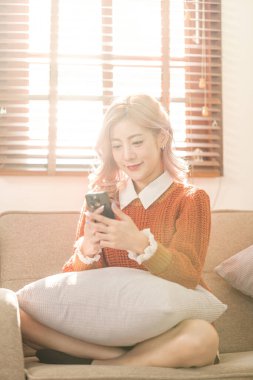 Odadaki kanepede oturan Asyalı genç bir kadın akıllı telefon kullanıyor. İnternetten alışveriş sipariş et. Yüksek kalite fotoğraf