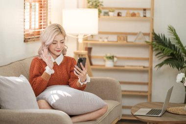 Odadaki kanepede oturan Asyalı genç bir kadın akıllı telefon kullanıyor. İnternetten alışveriş sipariş et. Yüksek kalite fotoğraf