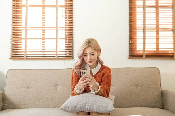 Odadaki kanepede oturan Asyalı genç bir kadın akıllı telefon kullanıyor. İnternetten alışveriş sipariş et. Yüksek kalite fotoğraf