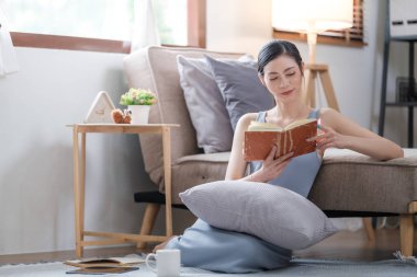 Evdeki kanepede kitap okuyan mutlu genç bir kadın. Yaşam tarzı, serbest çalışma, rahatlama ve oturma odasında kahve içme. Yaşam tarzı kavramı. Yüksek kalite fotoğraf