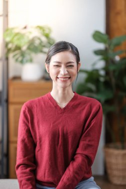 Güzel gülümseyen genç Asyalı kız oturma odasında sıradan kırmızı tişörtler giyiyor, gülümsüyor ve mutlu.