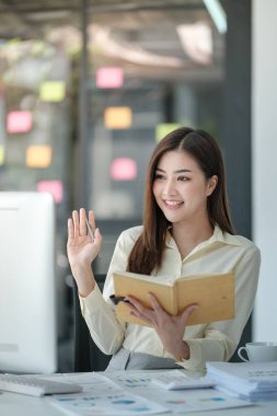 Çekici bir kadının masada oturup bilgisayarda çalışırken gülümsediği, elinde kitap tutan, elinde kahve tutan sarı tişörtlü bir kadının fotoğrafı. Yüksek kalite fotoğraf