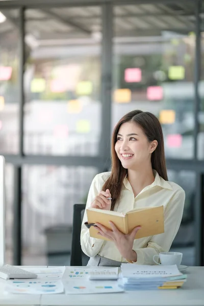 Çekici bir kadının masada oturup bilgisayarda çalışırken gülümsediği, elinde kitap tutan, elinde kahve tutan sarı tişörtlü bir kadının fotoğrafı. Yüksek kalite fotoğraf