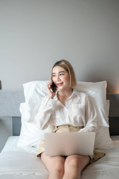 Cheerful asian woman Having Phone Conversation Using Laptop Sitting In Bed At Home. High quality photo