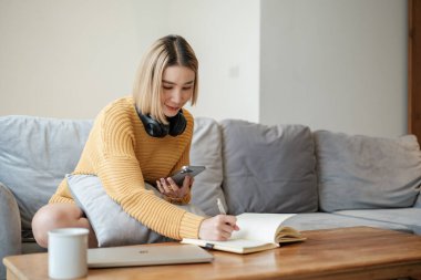 Asyalı bir kadın akıllı bir telefon kullanıyor ve gülen yüzlü bir kitap yazıyor, evde çalışıyor, ev hayatı tarzında, evde kalıyor. Yüksek kalite fotoğraf