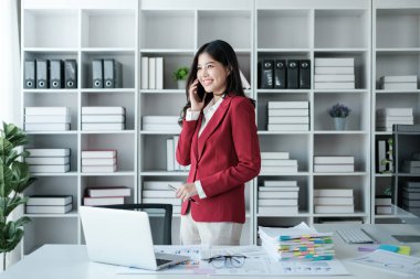 Güzel Asyalı iş kadını dizüstü bilgisayarda çalışıyor ve telefonla konuşuyor. Modern ofiste başarılı iş sonuçları kırmızı gömlek giyer. Yüksek kalite fotoğraf