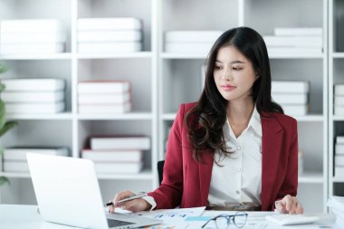 Güzel Asyalı iş kadını dizüstü bilgisayar üzerinde çalışıyor ve istatistik veri grafikleri ve grafiklerinden finansal hesap makinesi ile hesap yapıyor. Modern ofiste başarılı iş sonuçları kırmızı gömlek giyer. Yüksek kalite
