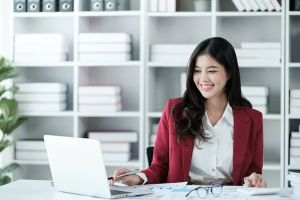 Güzel Asyalı iş kadını dizüstü bilgisayar üzerinde çalışıyor ve istatistik veri grafikleri ve grafiklerinden finansal hesap makinesi ile hesap yapıyor. Modern ofiste başarılı iş sonuçları kırmızı gömlek giyer. Yüksek kalite