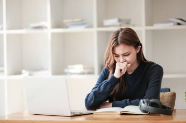 Stresli, beyaz tenli güzel bir kız öğrenci uzaktan çalışıyor. Evde dizüstü bilgisayar ve not defteriyle bir masada oturuyor ve bir video konferansı izliyor. Yüksek kalite fotoğraf