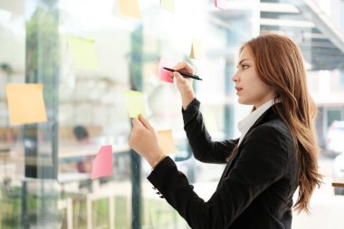 Güzel bayan çalışanlar renkli yapışkan notlar yönetici listesine yazarlar, konsantre çift ırklı kadınlar başlangıç beyin fırtınası planı üzerinde çalışırlar cam duvardaki çıkartmalar üzerinde işbirliği yaparlar. Yüksek kalite fotoğraf