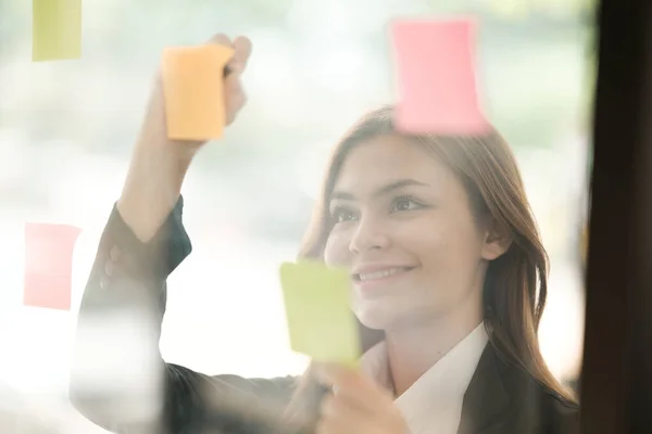 Beautiful female employee write down on colorful sticky notes manage list, concentrated biracial woman work on startup brainstorm collaborate plan on stickers on glass wall. High quality photo