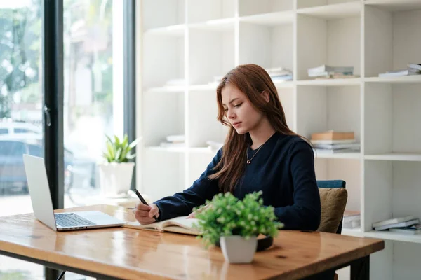 stock image A beautiful caucasian female student is studying in college remotely. She is sitting at a table at home with a laptop and a notepad and concentrated is watching a video conference lesson. High quality